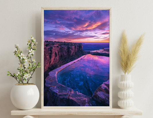 A stunning sunset view of a curved cliff with vibrant orange and purple hues reflecting on a pool of water and the distant horizon over the sea.
