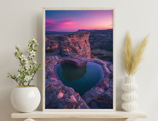 Heart-shaped lake reflecting mountains and pink-hued sky at sunset amidst rugged sandstone formations in a desert landscape.
