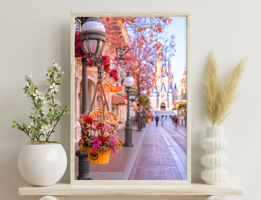 A festive autumn street scene at a theme park, adorned with colorful fall foliage, hanging flower baskets, jack-o'-lanterns, and the iconic castle in the background, creating a magical and vibrant atmosphere.
